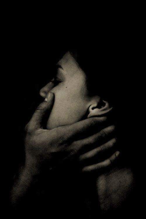 Close-up of a person in shadow with a hand over their mouth, conveying silence or suppression, in low light.